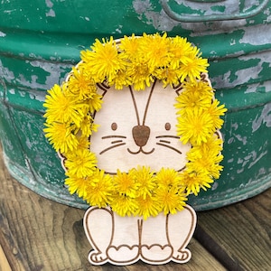 Może przedstawiać: Drewniane rzemiosło w kształcie lwa z żółtą koroną z kwiatów mniszka lekarskiego. Lew ma prosty, kreskówkowy design z wytrawionymi detalami. Tło obejmuje zielone wiadro i drewnianą powierzchnię.