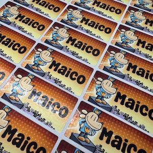 May include: Rectangular stickers with the name "Maico" in bold black letters. Each sticker features a cartoon dog character in a blue outfit and a crown. The background has a gradient of orange and yellow.