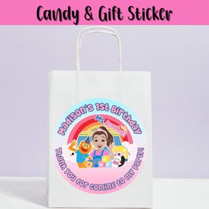 May include: White paper gift bag with a round sticker. The sticker says "Madison's 1st Birthday" with a rainbow and cartoon characters. Text on the sticker reads "Thank you for coming to my party!" and "Ms. Rachel". The text "Candy & Gift Sticker" is at the top.