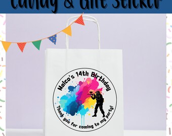 May include: White paper gift bag with a circular sticker that reads "Maico's 14th Birthday" with a paintball player graphic. The sticker has a colorful paint splatter design and the text "Thank you for coming to my party!" The bag has a white handle and a banner of colorful flags.