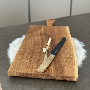 Puede incluir: Una tabla de cortar de madera r&uacute;stica grande con un borde vivo y un asa. Un cuchillo negro dentado con mango de madera descansa sobre la tabla.