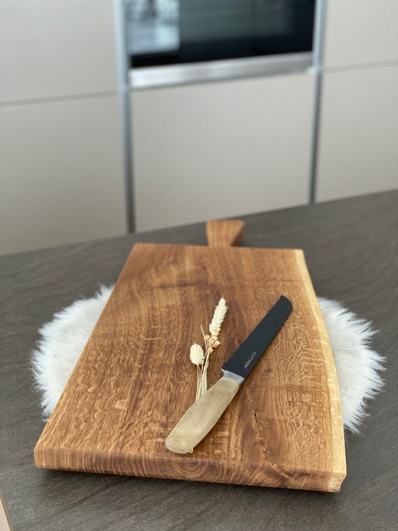 Puede incluir: Una tabla de cortar de madera r&uacute;stica grande con un borde vivo y un asa. Un cuchillo negro dentado con mango de madera descansa sobre la tabla.