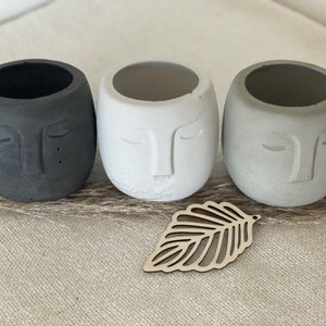 Könnte beinhalten: Drei kleine Beton-Pflanztöpfe in Form von Gesichtern mit geschlossenen Augen. Die Töpfe sind in Schwarz, Weiß und Grau. Im Vordergrund befindet sich ein Holz-Blatt-Ausschnitt.