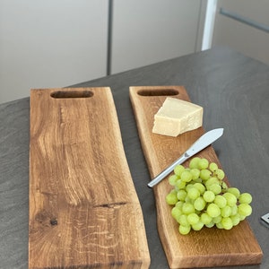 Puede incluir: Dos tablas de servir de madera con veta de madera natural y un asa en cada extremo. Una tabla tiene una cu&ntilde;a de queso y un racimo de uvas verdes. Un cuchillo plateado est&aacute; sobre la tabla.