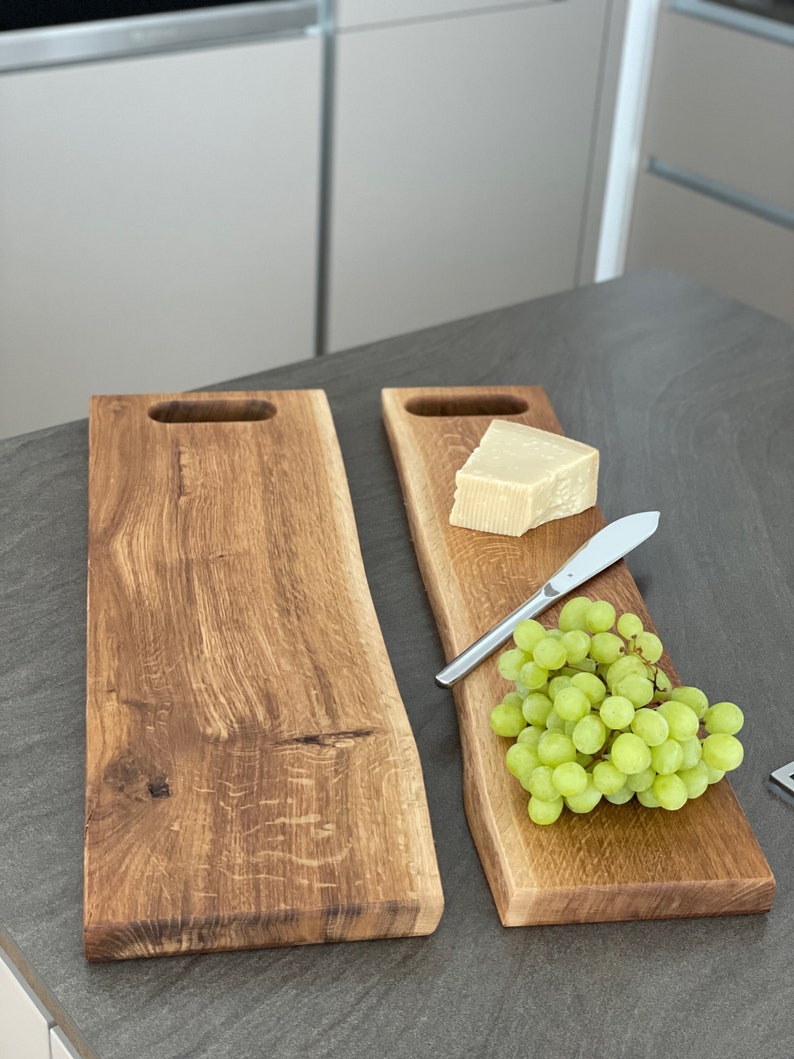 Puede incluir: Dos tablas de servir de madera con veta de madera natural y un asa en cada extremo. Una tabla tiene una cu&ntilde;a de queso y un racimo de uvas verdes. Un cuchillo plateado est&aacute; sobre la tabla.