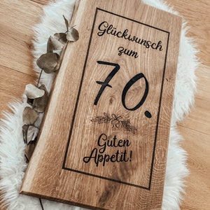 Puede incluir: Una tabla de cortar de madera con un dise&ntilde;o grabado en negro que dice "Gl&uuml;ckwunsch zum 70. Guten Appetit!" La tabla tiene un asa en la parte superior.