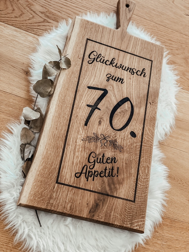 Puede incluir: Una tabla de cortar de madera con un dise&ntilde;o grabado en negro que dice "Gl&uuml;ckwunsch zum 70. Guten Appetit!" La tabla tiene un asa en la parte superior.
