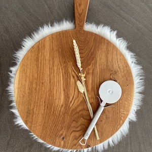 Puede incluir: Una tabla de servir redonda de madera con un asa, hecha de madera marr&oacute;n claro con un patr&oacute;n de veta natural. La tabla est&aacute; rodeada por un ribete de piel sint&eacute;tica blanca. Un cortador de pizza plateado est&aacute; sobre la tabla.