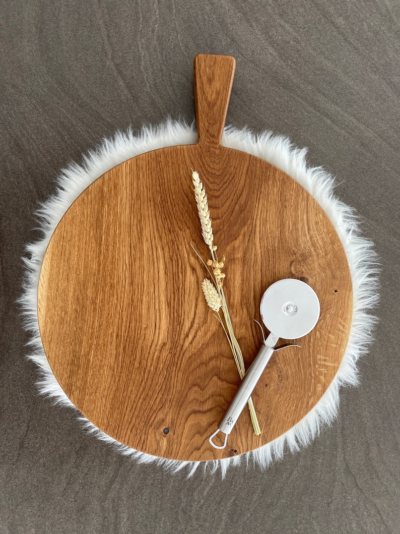 Puede incluir: Una tabla de servir redonda de madera con un asa, hecha de madera marr&oacute;n claro con un patr&oacute;n de veta natural. La tabla est&aacute; rodeada por un ribete de piel sint&eacute;tica blanca. Un cortador de pizza plateado est&aacute; sobre la tabla.