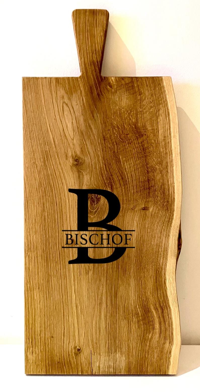 Puede incluir: Una tabla de cortar de madera grande, rectangular, con un borde vivo y un asa. La tabla est&aacute; hecha de madera marr&oacute;n claro con un acabado natural. La tabla tiene la letra "B" y el nombre "BISCHOF" grabados.