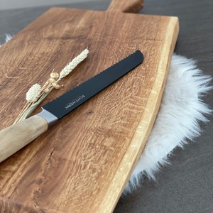 Puede incluir: Una tabla de cortar de madera grande, rectangular, con un borde vivo y un acabado de madera natural. Un cuchillo de pan negro serrado con mango de madera descansa sobre la tabla. La tabla est&aacute; sobre una alfombra de piel sint&eacute;tica blanca.