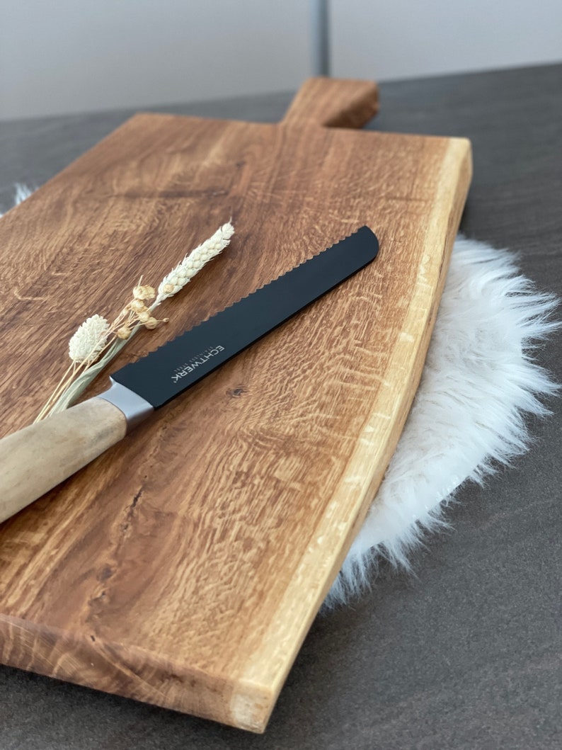 Puede incluir: Una tabla de cortar de madera grande, rectangular, con un borde vivo y un acabado de madera natural. Un cuchillo de pan negro serrado con mango de madera descansa sobre la tabla. La tabla est&aacute; sobre una alfombra de piel sint&eacute;tica blanca.