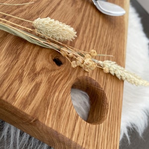 Puede incluir: Una tabla de servir de madera con un asa redonda recortada. La tabla es de madera marr&oacute;n claro con un patr&oacute;n de veta natural. Flores secas se colocan en la tabla.