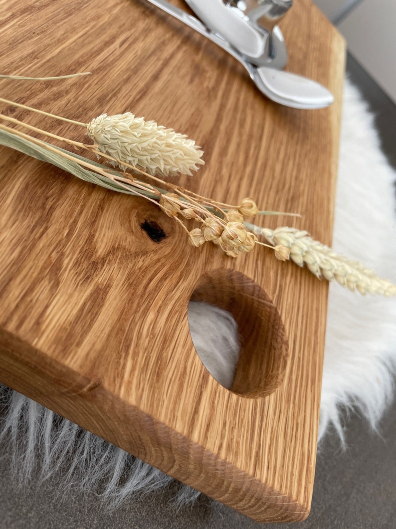 Puede incluir: Una tabla de servir de madera con un asa redonda recortada. La tabla es de madera marr&oacute;n claro con un patr&oacute;n de veta natural. Flores secas se colocan en la tabla.