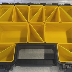 Może przedstawiać: Czarny i żółty organizer na części z wieloma przegródkami. Organizer ma czarny uchwyt i przezroczystą pokrywę. Wnętrze posiada kilka żółtych pojemników, niektóre z kątowymi przegrodami, przeznaczonych do organizacji drobnych przedmiotów.