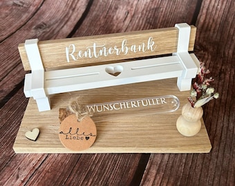 May include: A wooden bench with a white seat and back, labeled "Rentnerbank" with a heart cutout. A small glass tube with the text "Wunscherfueller" is on the bench, and a brown tag with the text "alles Liebe" is on the base.