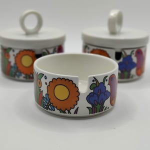 Puede incluir: Un juego de tres cuencos de cerámica blanca con tapas. Los cuencos tienen un diseño floral y de pájaros en colores brillantes, incluyendo naranja, amarillo, azul, verde y rojo.