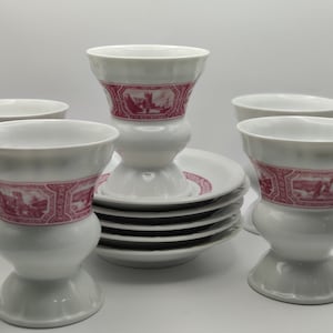 May include: Set of six white porcelain coffee cups with pink floral designs. The cups have a wide base and a narrow top. The cups are stacked on top of each other.