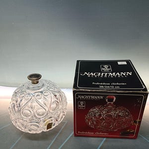 Peut inclure: Un bonbonnière en verre transparent avec un couvercle, ornée d'un motif décoratif de cercles et de volutes. Le plat est étiqueté "NACHTMANN BLEIKRISTALL Pralinédose >Sinfonie< 38/24/12 cm".