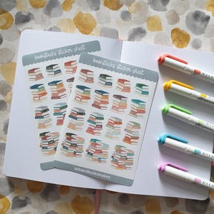 May include: Two sticker sheets with stacks of books, labelled "bookstacks sticker sheet," are on a white notebook. The stickers feature colourful book illustrations. Five white pens with coloured tips are also visible.