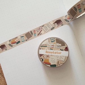 May include: A roll of washi tape with a bookish design. The tape displays illustrations of books, spectacles, and floral motifs in a repeating pattern. The roll is labelled "Annotator" and is 1.5cm x 10m.