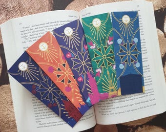 Puede incluir: Cinco marcapáginas coloridos con un diseño de ancla dorada y patrones florales. Los marcapáginas están dispuestos en forma de abanico sobre un libro.