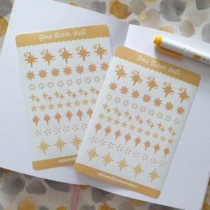 May include: Two sticker sheets with a light yellow border and a white background. The sheets are filled with various star designs in shades of yellow and orange. The text "Stars sticker sheet" is at the top, and "ashandbooksmakes" is at the bottom.