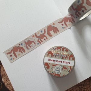 May include: A roll of washi tape and a strip of extended tape featuring a repeating pattern of reddish-brown creatures, geometric shapes, and small stars. The label on the roll reads 2.5cm x 10m washi tape, Rocky Save Stars, and Ash & Books makes.