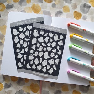 May include: Two sticker sheets with white ghost illustrations on a dark gray background, labeled "ghosties sticker sheet." The stickers are on a white notebook, with colorful pens to the right. The background is a watercolor pattern.