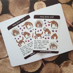 May include: Two white sticker sheets labelled rocky sticker sheet and ashandbooksmakes featuring brown spider-like creatures, colourful stars, and text including WHY STUPID. QUESTION? GRACE ROCKY SAVE STARS AMAZE. AMAZE. AMAZE ROCKY HATE MARK DIRTY DIRTY DIRTY. WHY SO MESSY.
