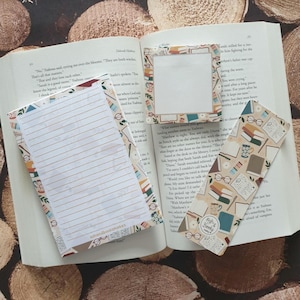 May include: A set of three bookish themed stationery items: a notepad with a floral and glasses pattern, a bookmark with a similar pattern, and a blank notecard with a brown frame. The bookmark has the text "Read & Repeat" on it.