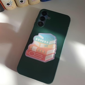 May include: A dark green phone case with a sticker of a stack of books. The sticker shows a stack of books in red, orange, yellow, and blue. The books have decorative details and are outlined in pink.