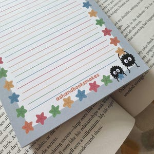 May include: A lined notepad with a light blue border adorned with colorful stars. The notepad features rainbow-colored lines and two cartoon soot sprites. The text "ashandbooksmakes" is printed on the notepad.