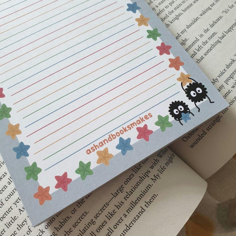 May include: A lined notepad with a light blue border adorned with colorful stars. The notepad features rainbow-colored lines and two cartoon soot sprites. The text "ashandbooksmakes" is printed on the notepad.