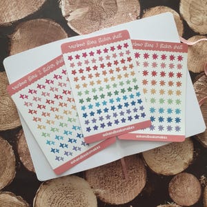 May include: Three rectangular sticker sheets featuring rows of small, colorful stars in rainbow gradients. Each sheet is labeled with text including rainbow stars, sticker sheet, and ashandbooksmakes, printed on a white background with pink borders.