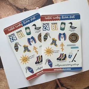 May include: Two sticker sheets with red and blue borders, displaying illustrations and text. Designs include a beer can, a duck, keys, hockey skates, and the text "heated rivalry sticker sheet".