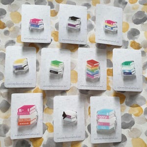 May include: A selection of book-shaped pins displayed on white cards. The pins depict stacks of books in diverse colours, such as pink, blue, green, and yellow. The text "ashandbooksmakes" is printed at the base of each card.