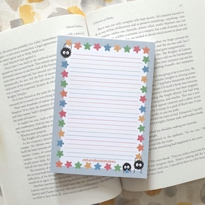 May include: A lined notepad with a colorful star border and whimsical monster illustrations. The notepad has the text "ashandbooksmakes" at the bottom. The notepad is resting on top of an open book.