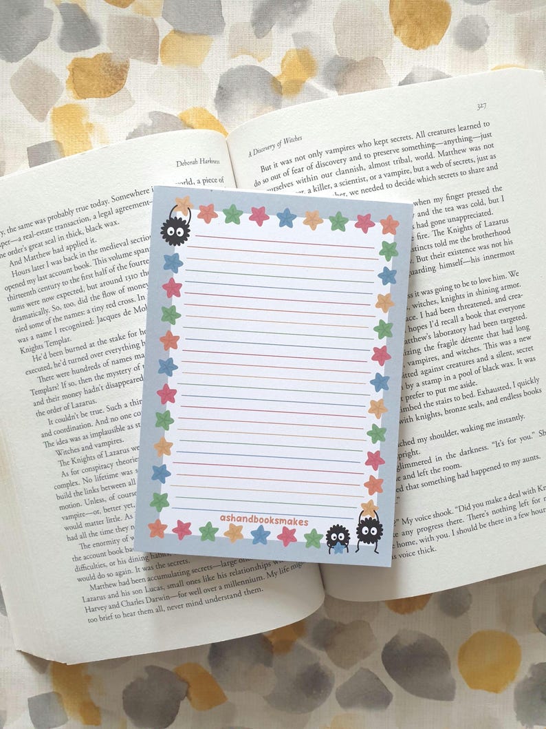 May include: A lined notepad with a colorful star border and whimsical monster illustrations. The notepad has the text "ashandbooksmakes" at the bottom. The notepad is resting on top of an open book.