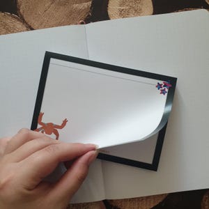 May include: A rectangular notepad with a black border and white pages. The top page features a small orange crab illustration on the left and a cluster of three blue and red star shapes on the right. The bottom edge shows partially visible text.