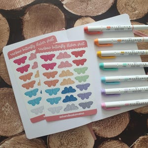 May include: Two white sticker sheets labeled rainbow butterfly sticker sheet with rows of colorful butterfly stickers, alongside seven double-ended Zebra Mildliner markers in various colors, all resting on an open white notebook.