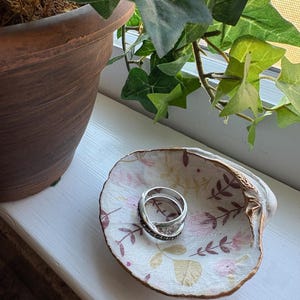 May include: A decorative seashell dish with a floral pattern in pink and beige, holding two silver rings. The dish sits on a white windowsill, with a potted plant and green ivy in the background.