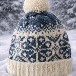 May include: A knitted winter hat with a cream-coloured pom-pom and brim. The hat features a blue and cream snowflake pattern. The hat is in the snow, with a snowy forest in the background.