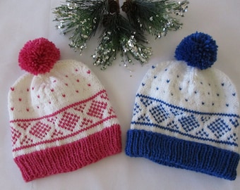 Puede incluir: Dos gorros de invierno de punto con pompones. Un gorro es blanco con un patrón rosa y blanco y un pompón rosa. El otro gorro es blanco con un patrón azul y blanco y un pompón azul.