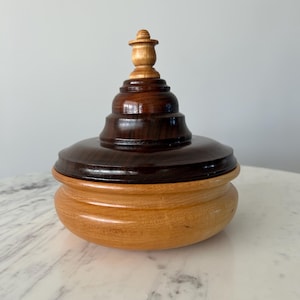May include: A wooden box with a lid, featuring a dark brown top and a light brown base. The lid has a small, light brown knob on top.