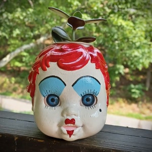 May include: A red, white, and blue ceramic planter shaped like a doll's face with blue eyes, red lips, and a heart-shaped nose. The planter is decorated with red hair and has a small green plant growing out of the top.