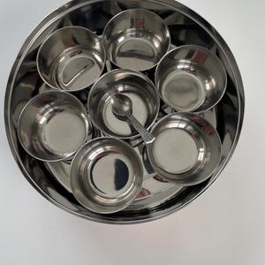 May include: A round stainless steel spice container with seven individual bowls and a small spoon.