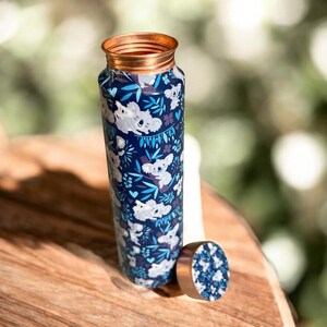 May include: A stainless steel water bottle with a copper rim and a screw-on lid. The bottle is navy blue and covered in a pattern of white koalas, blue leaves, and floral accents. The lid is off to the side.