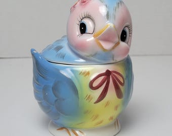 Könnte beinhalten: Ein blauer und gelber Keramikvogel mit rosa Kopf und roter Schleife. Der Vogel hat einen Deckel und ist eine Zuckerdose.