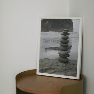 Puede incluir: Una fotografía enmarcada en blanco de una pila de piedras en una playa con una isla verde en el fondo. La fotografía está sobre una mesa de madera con una base de metal dorado.